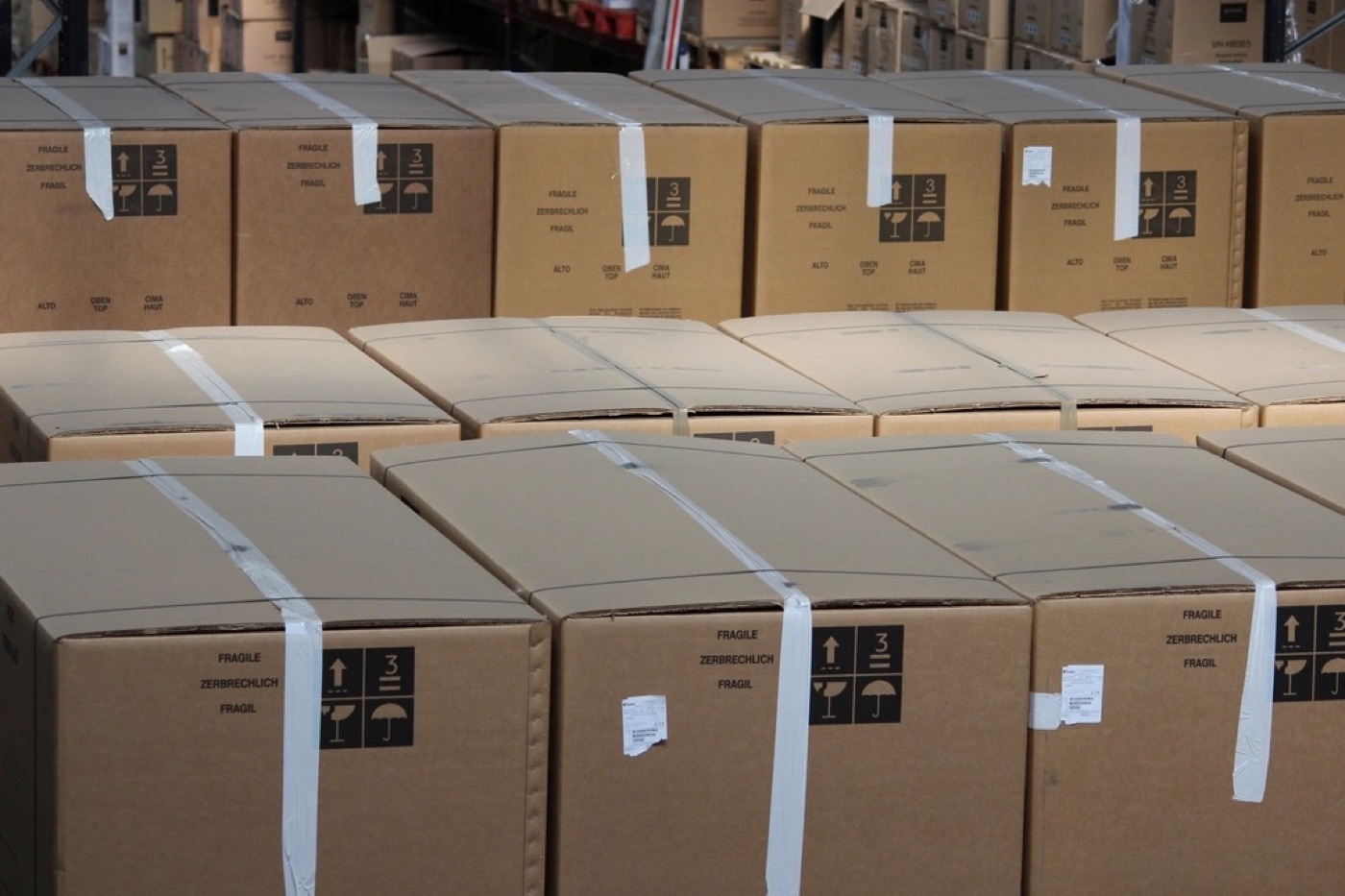 Rows of taped cardboard boxes stacked on a warehouse floor, each marked with fragile and this-way-up symbols