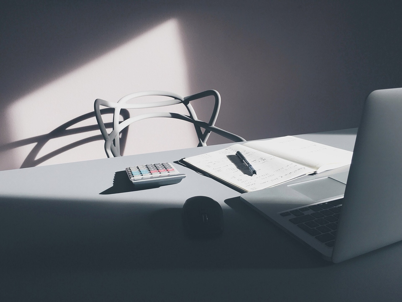 A silver laptop, an open handwritten notebook with a pen, a calculator and a mouse on a gray desk in hard window light