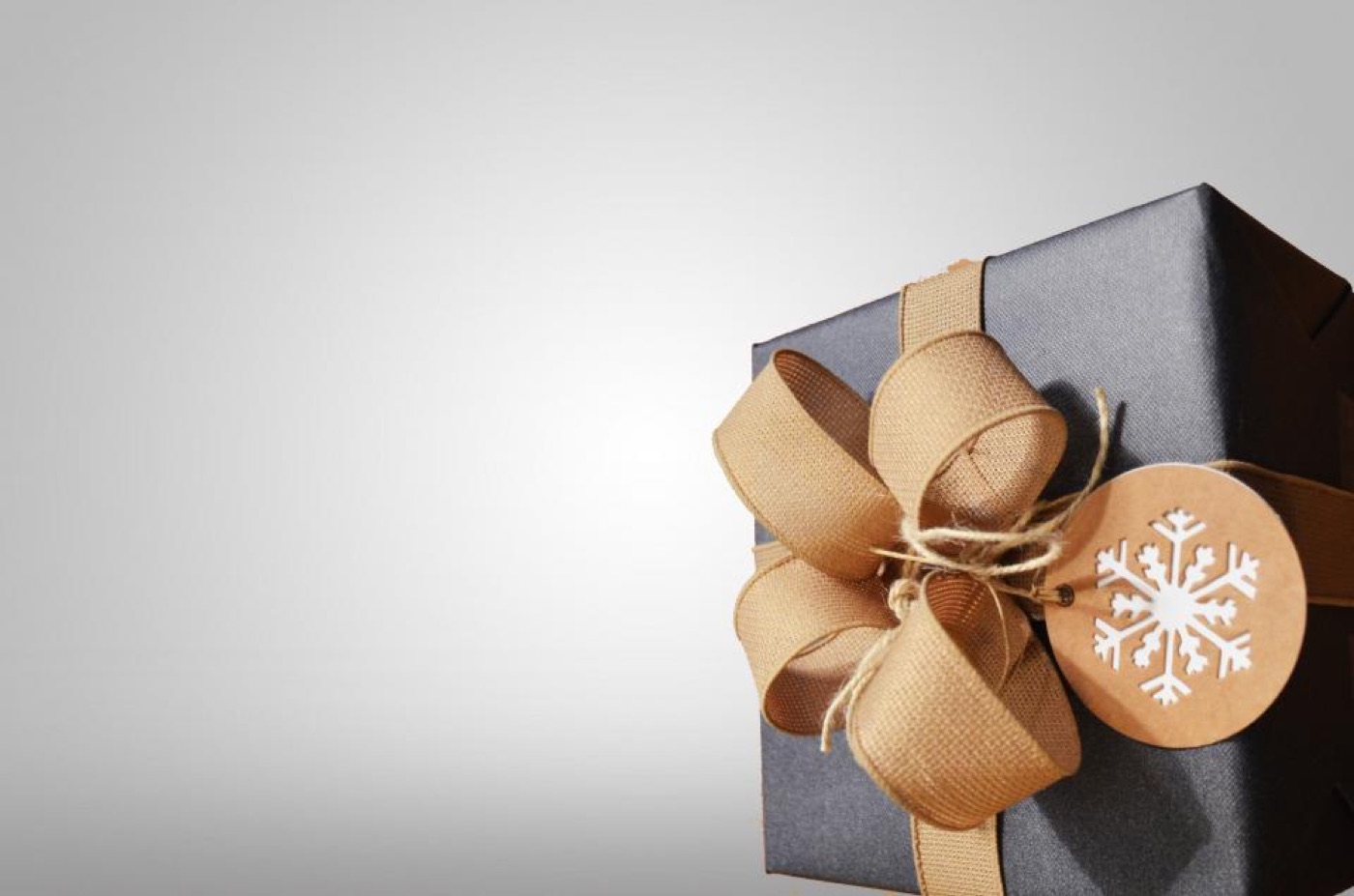A gift box wrapped in dark paper, tied with a wide tan ribbon bow and a round tag
