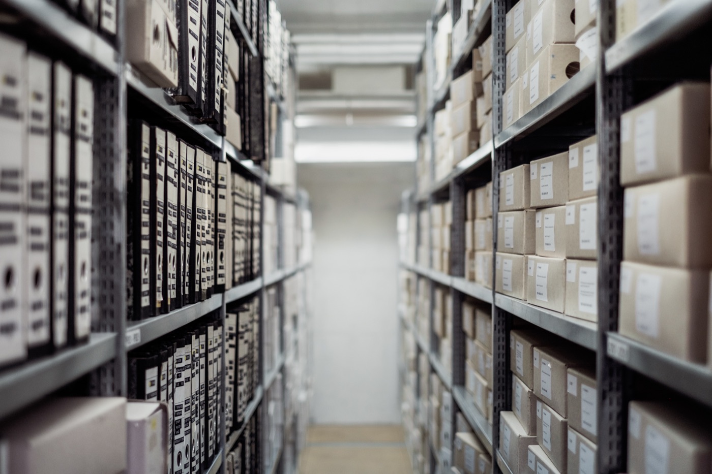 A narrow aisle between metal shelving racks stacked with cardboard storage boxes and black binders, leading to a bright end wall