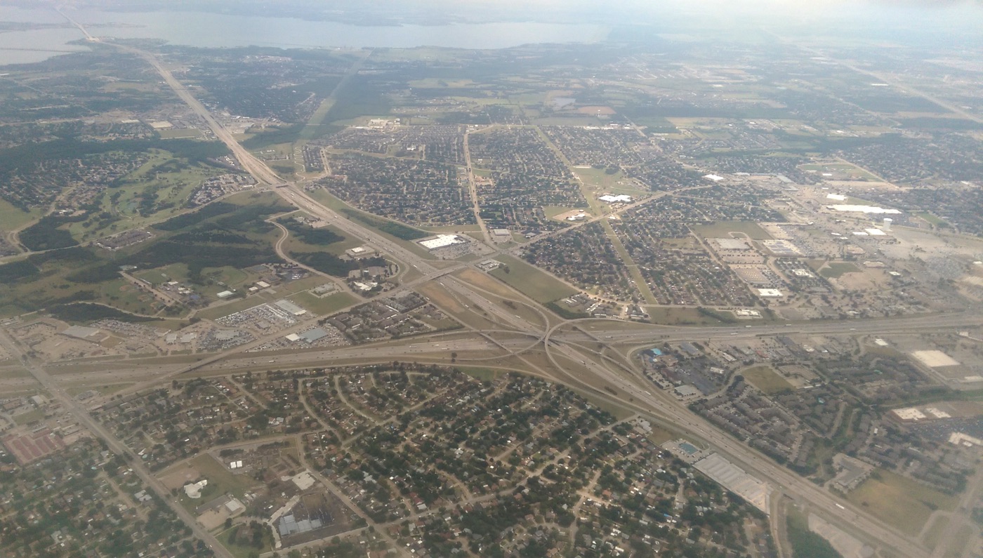 Aerial view of a freeway interchange where two major highways cross, with neighborhoods and shopping centers around it and a lake on the horizon