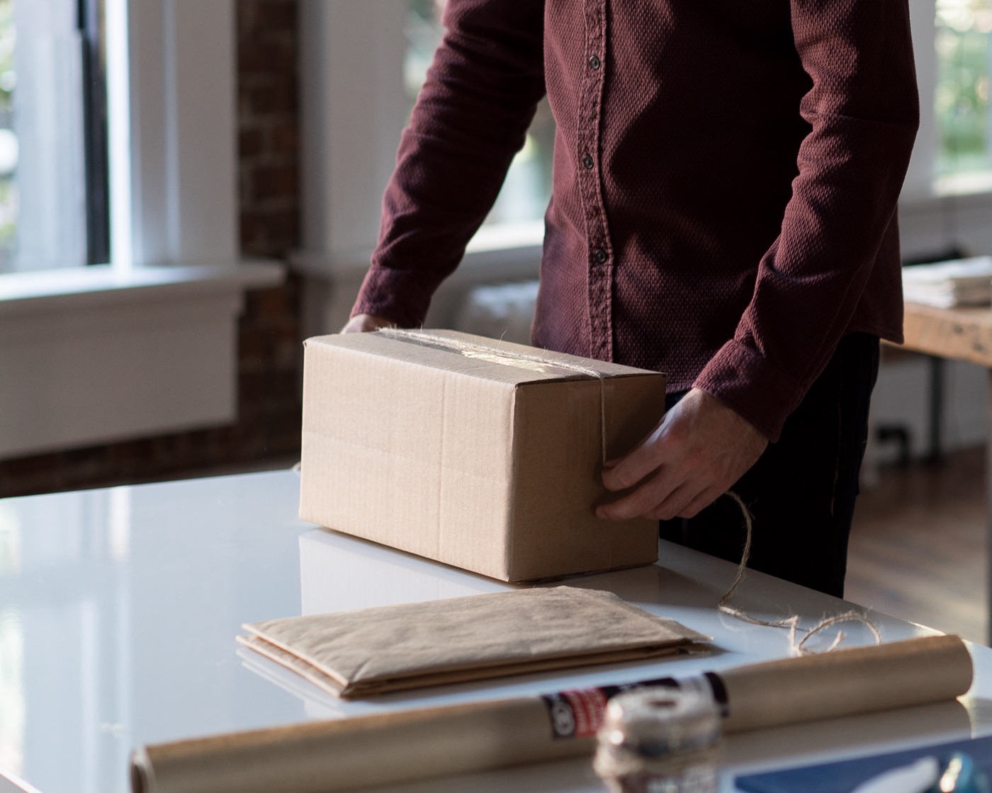 Person in a maroon button-up shirt, face out of frame, tying twine around a taped cardboard box on a white packing table, with a padded mailer and a roll of kraft paper beside it
