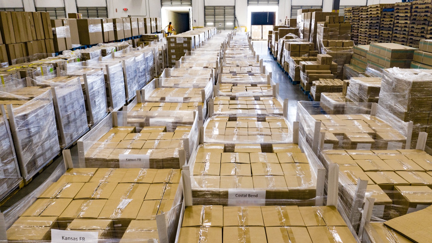 Pallets of taped cardboard cartons wrapped in clear stretch film, lined up in rows on a warehouse floor with dock doors at the far end